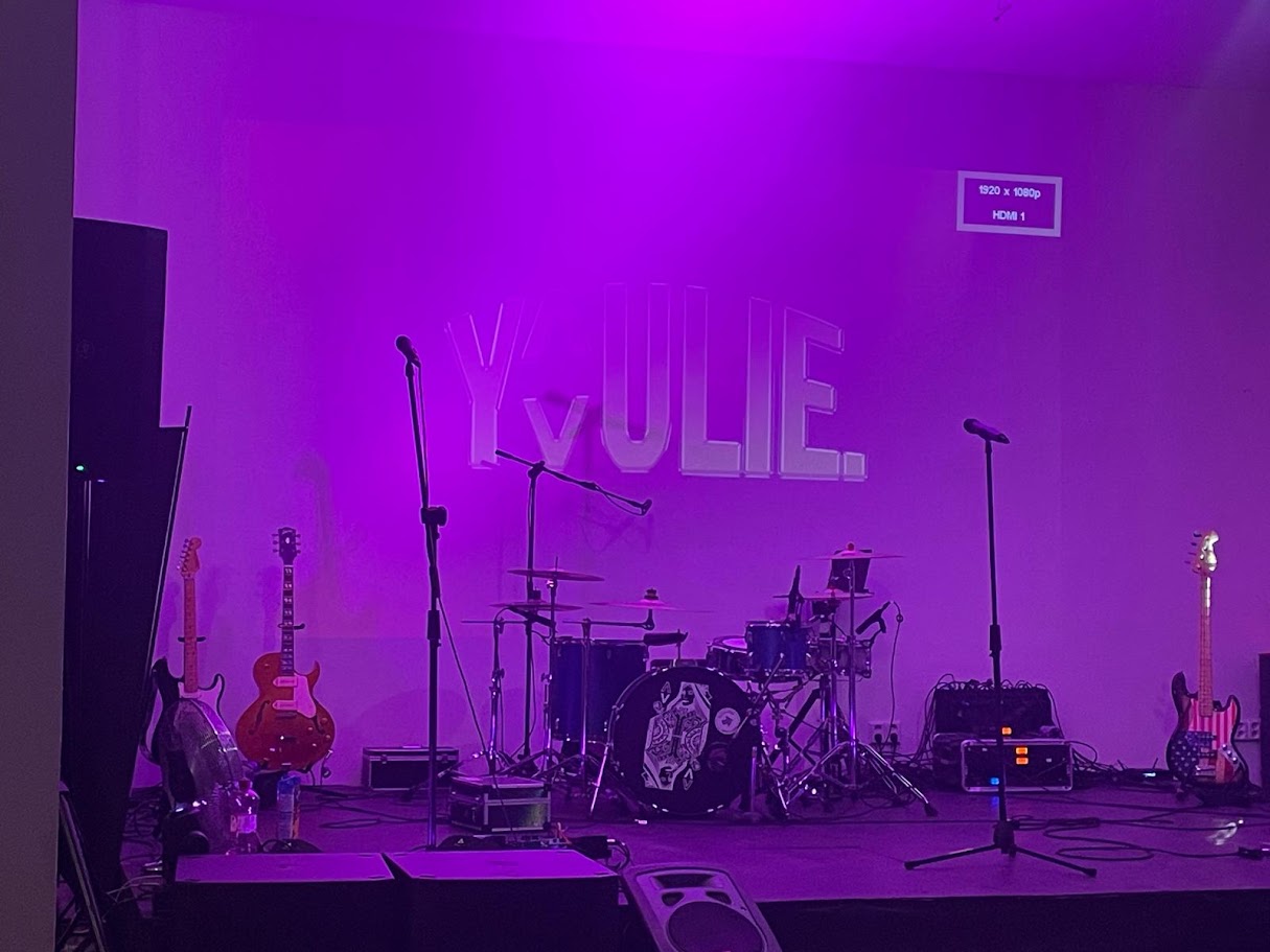 Připravená klubová stage s bicími, mikrofony a fialovým světlem.